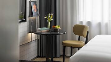 A modern hotel room with a round table, a chair, a bed, a small table with books and a vase of flowers, and a window with curtains.