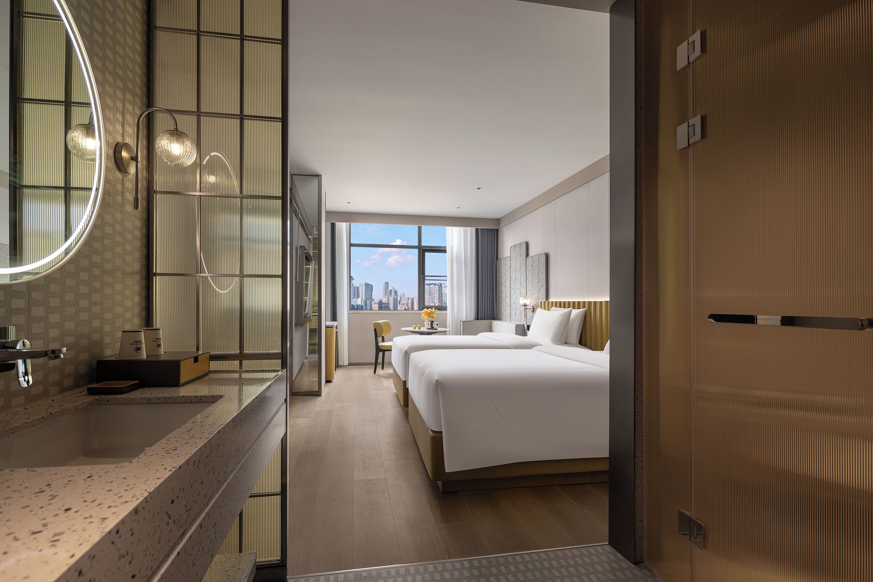 Een hotelkamer met een groot bed, een zithoek met een stoel en tafel, een badkamer met spiegel en wastafel, en uitzicht op de skyline van de stad.