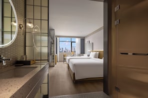 A hotel room with a large bed, a sitting area with a chair and table, a bathroom with a mirror and sink, and a view of the city skyline through the window.
