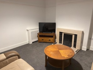 A living room with a fireplace, a television on a stand, and a round wooden table. - Central Peterborough Home, Garden, Parking, 2 Bed (Peterborough)