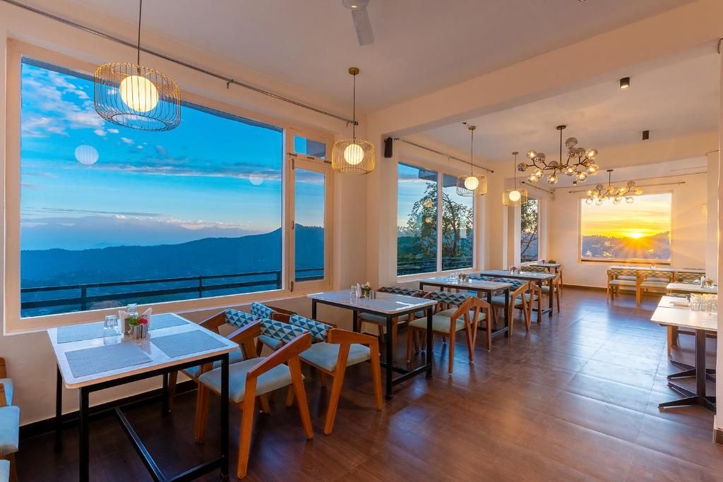 Comedor con mesas y sillas, ventanas amplias con vistas al cielo y al paisaje, e iluminación colgante.