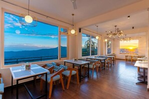 A dining area with tables and chairs, large windows with views of the sky and landscape, and pendant lighting. - Himalayas Resort by The Lake Hill (Nainital)