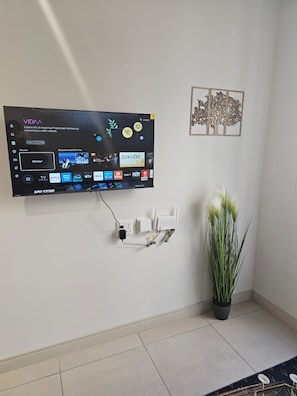 Ein an einer Wand befestigter Flachbildfernseher zeigt verschiedene Streaming-Dienste an, daneben steht eine Pflanze.