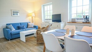 Een moderne woonkamer met een blauwe bank, een witte salontafel, een rieten fauteuil, en een eethoek met witte stoelen en ronde tafels.