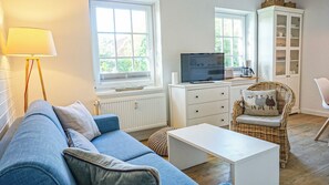 A living room with a sofa, a television, a coffee table, a wicker chair, and a cabinet. - Deichnest Vollerwiek Apartment: A Relaxing Getaway by the Sea (Vollerwiek)