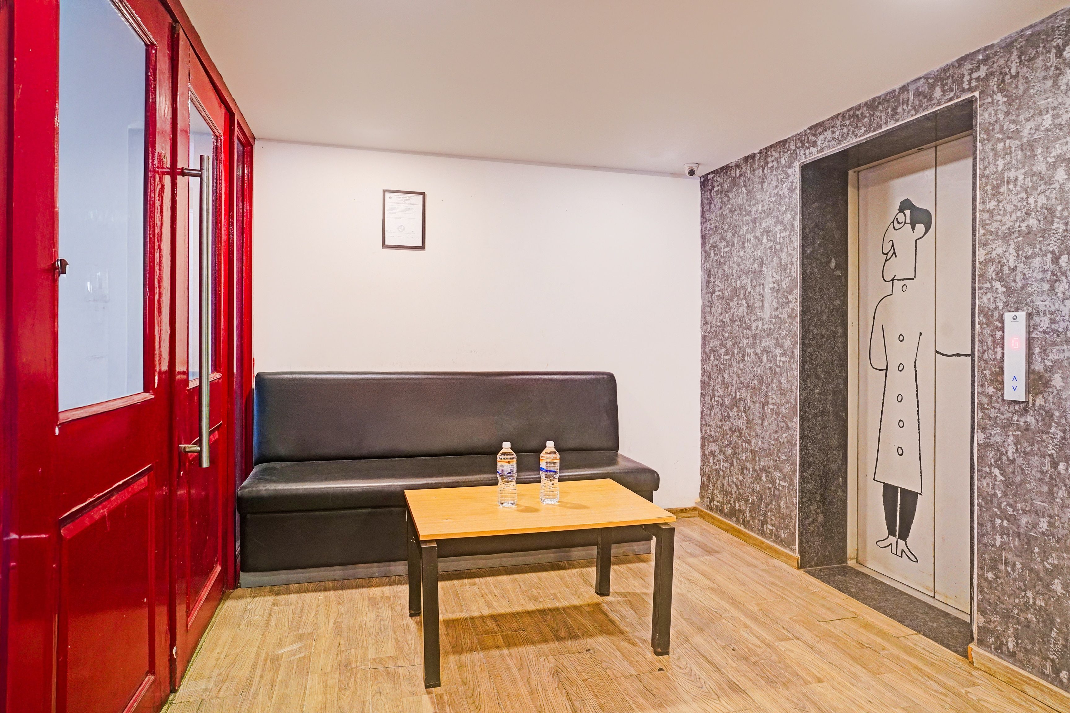 Une pièce avec une porte rouge, un canapé noir, une table en bois sur laquelle se trouvent deux bouteilles d’eau, et une caméra de surveillance fixée au mur.