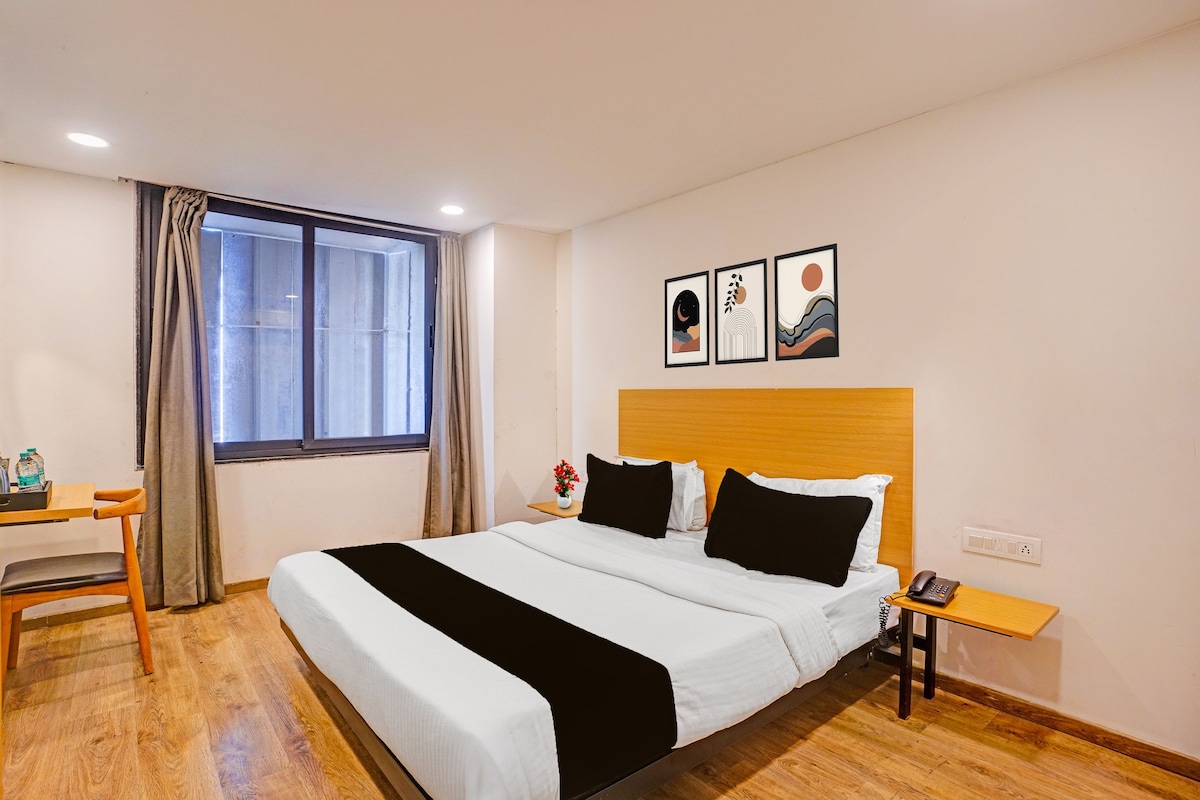 A hotel room with a bed, bedside table, a desk with a chair, a window with curtains, and wall art.