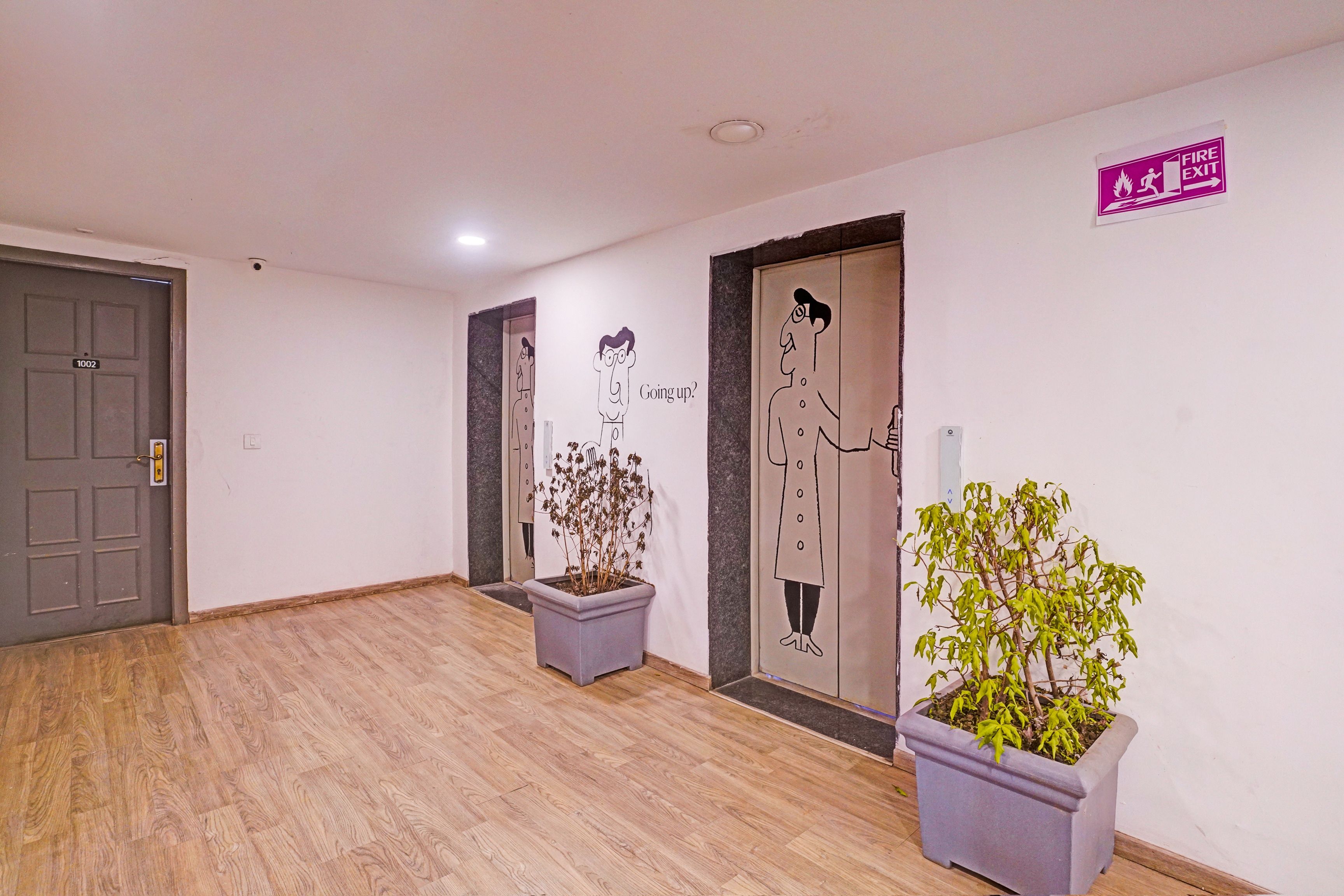 A hallway with a grey door labeled '1002', a fire exit sign, and two potted plants.