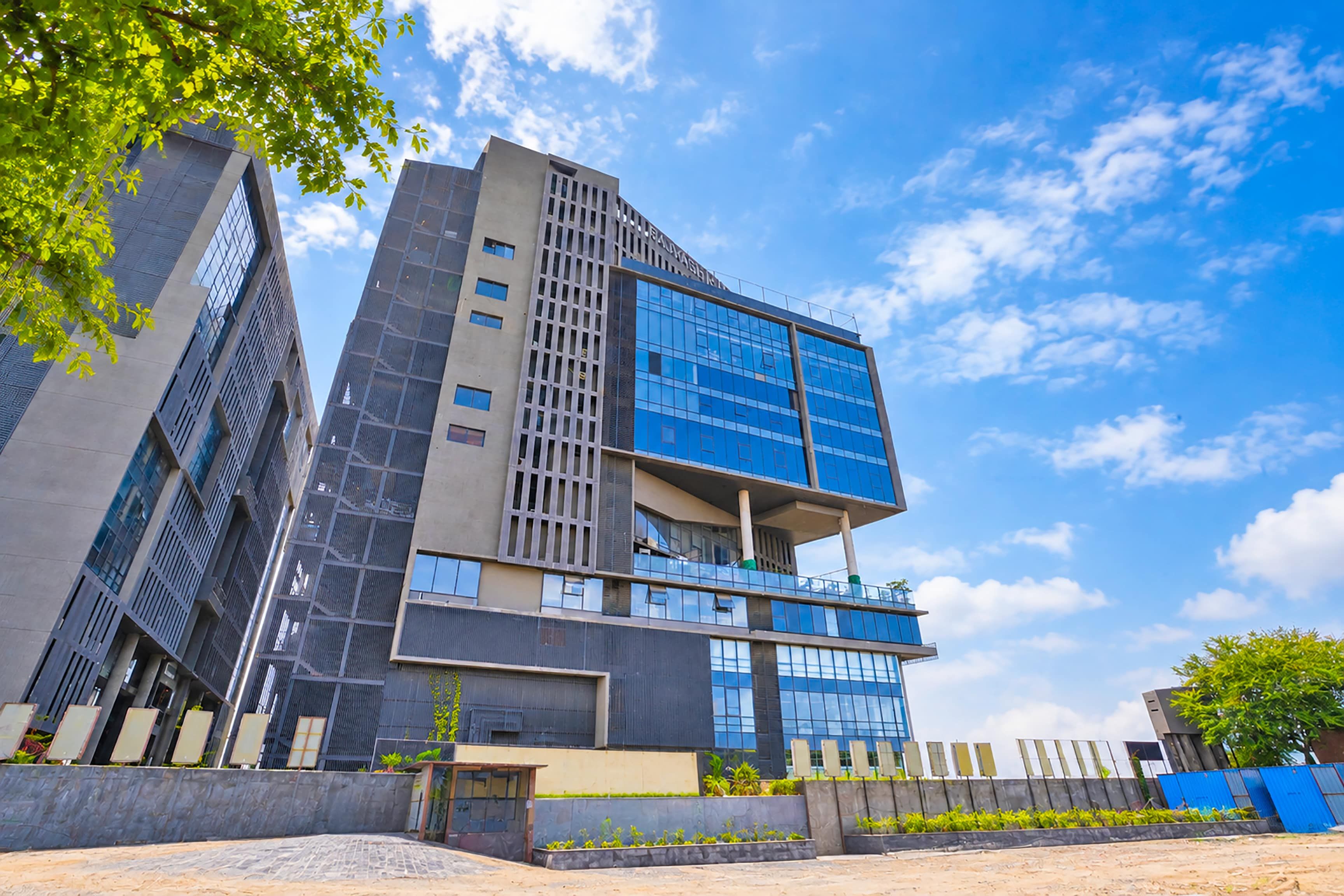 Un edificio moderno de varios pisos con fachada de vidrio y metal, rodeado de vegetación y un cielo despejado.