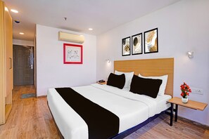 A hotel room with a bed, a bedside table, a nightstand, a wall-mounted air conditioner, a framed picture, and a wall-mounted light fixture. - Townhouse APMC Metro Station  (Ahmedabad)