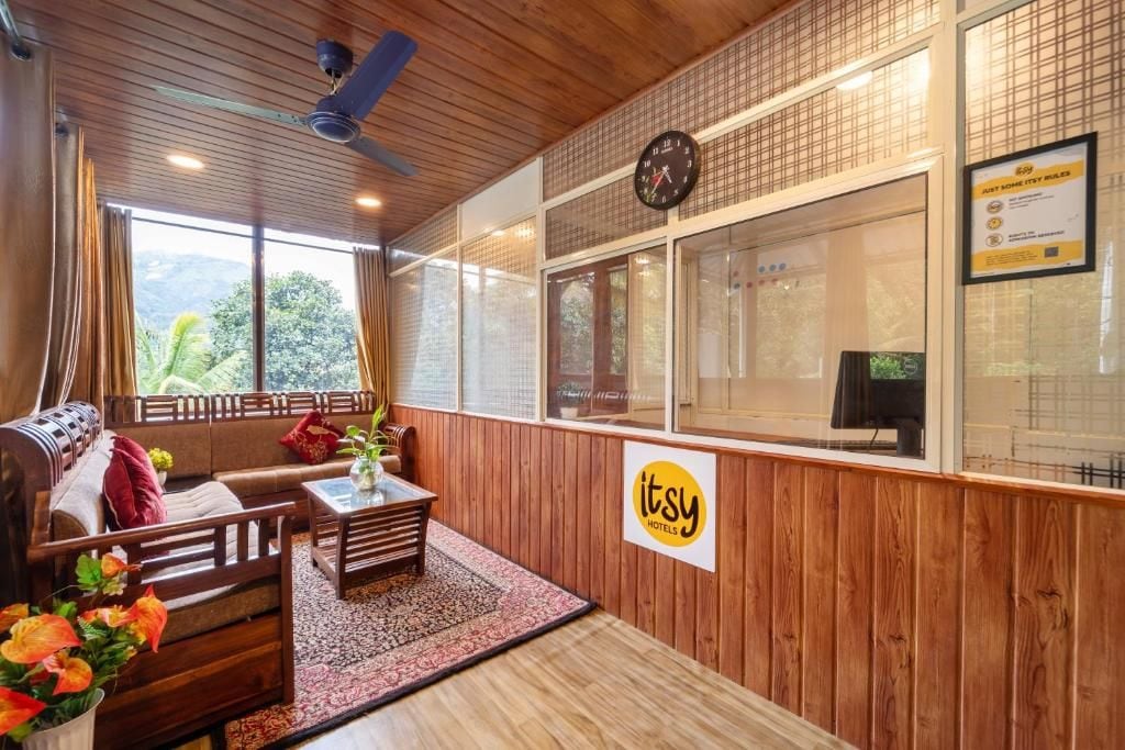 A hotel room with a wooden ceiling, a sofa, a coffee table, a clock, and a window with a view of greenery.