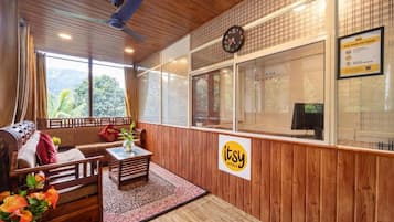 A hotel room with a wooden ceiling, a sofa, a coffee table, a clock, and a window with a view of greenery.