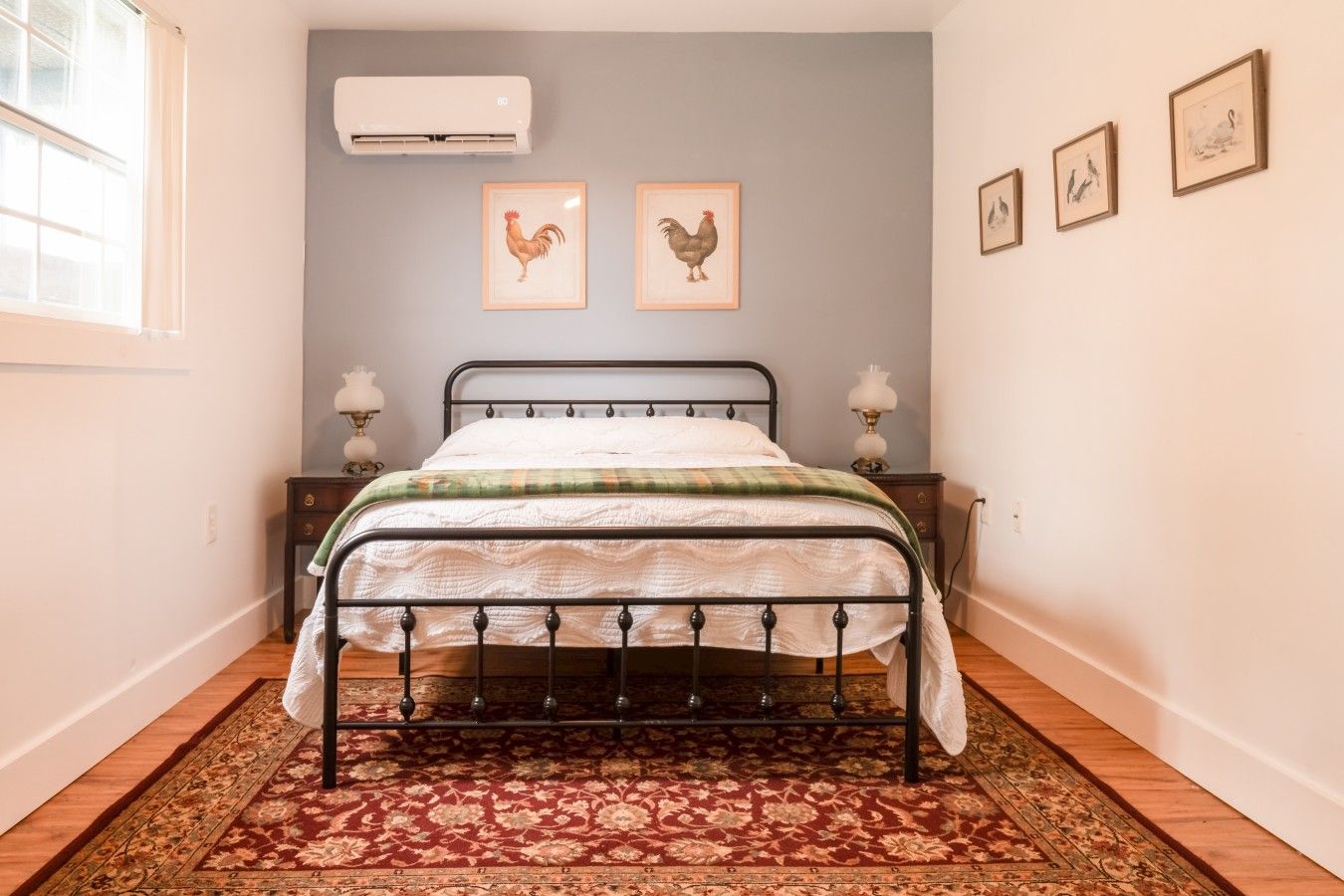 Een slaapkamer met een bed met metalen frame, een bedovertrek met patroon, een tapijt in rood en beige tinten, een wandairconditioning en ingelijste foto's aan de muur.