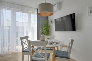 A modern living room with a round dining table, white chairs, a potted plant, and a flat-screen TV mounted on the wall. - Portovo 96 Apartment with Parking (Gdańsk)