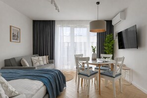 Una habitación de hotel moderna con cama, sofá, mesa de comedor, sillas, televisor y un ventanal con cortinas.
