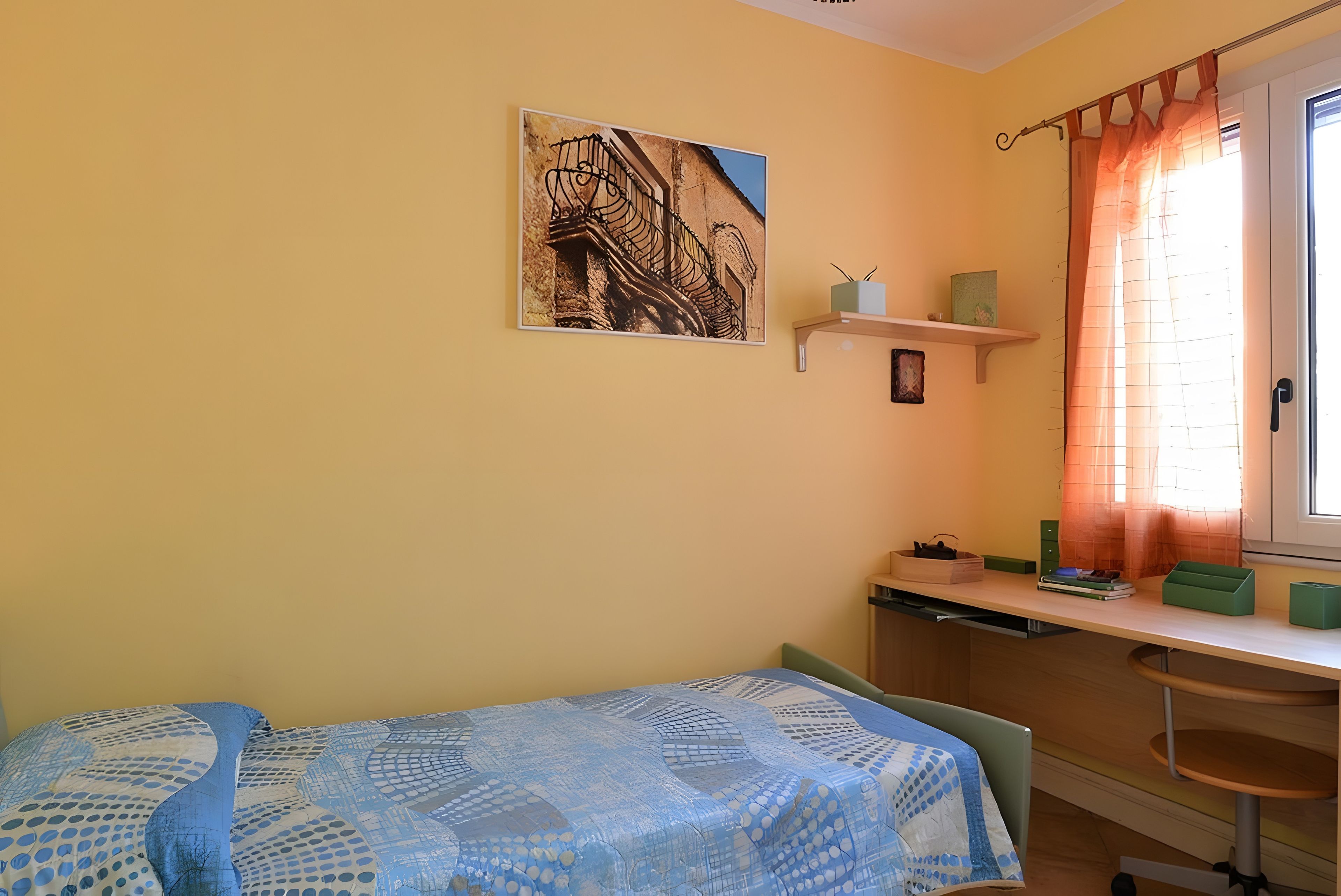 Een slaapkamer met een bed, een bureau, een stoel, een raam met gordijnen, en een wandplank met spullen.