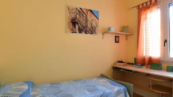 Een slaapkamer met een bed, een bureau, een stoel, een raam met gordijnen, en een wandplank met spullen.
