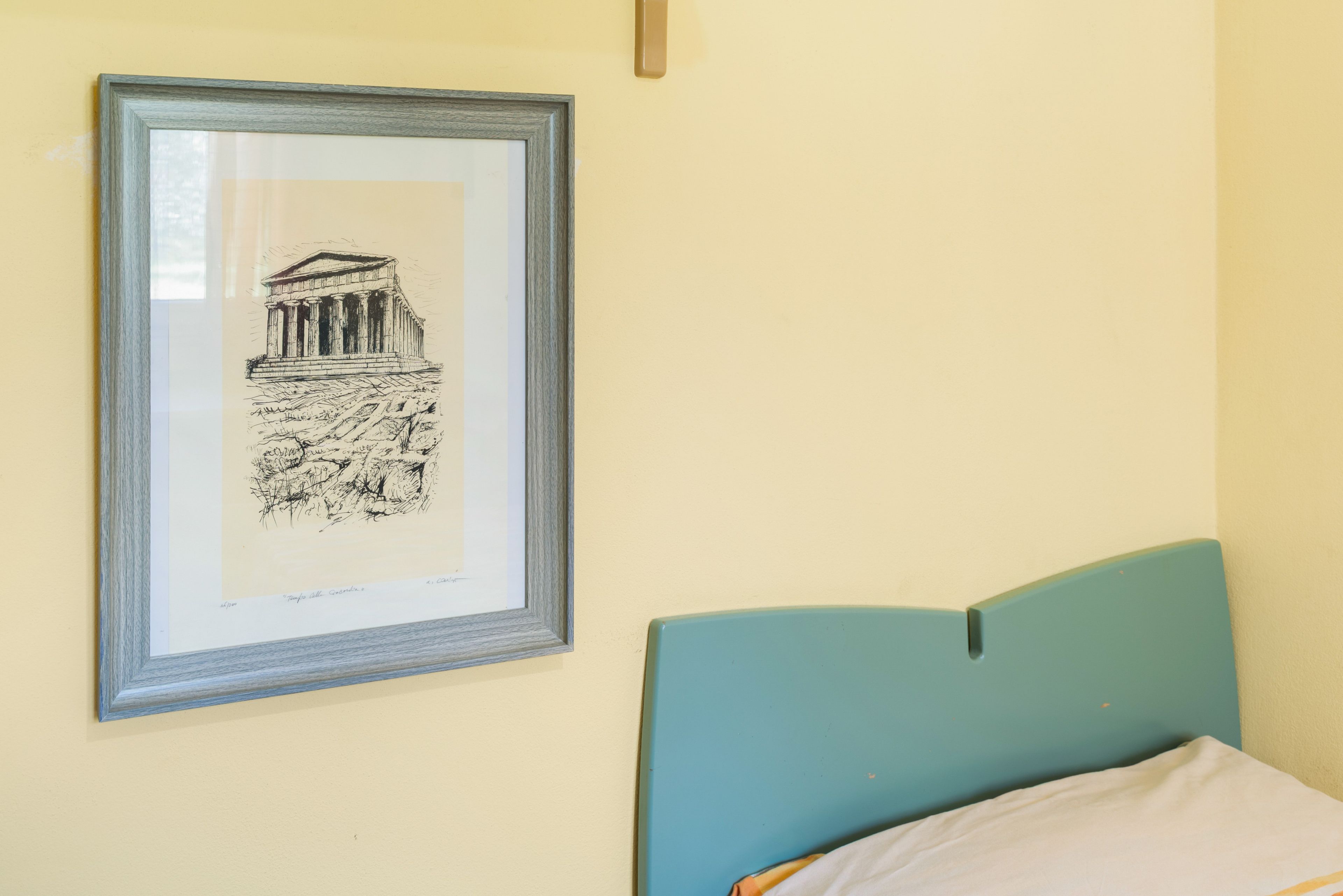 A framed black and white drawing of a classical building on a wall, a bed with a teal headboard, and a plain wall.