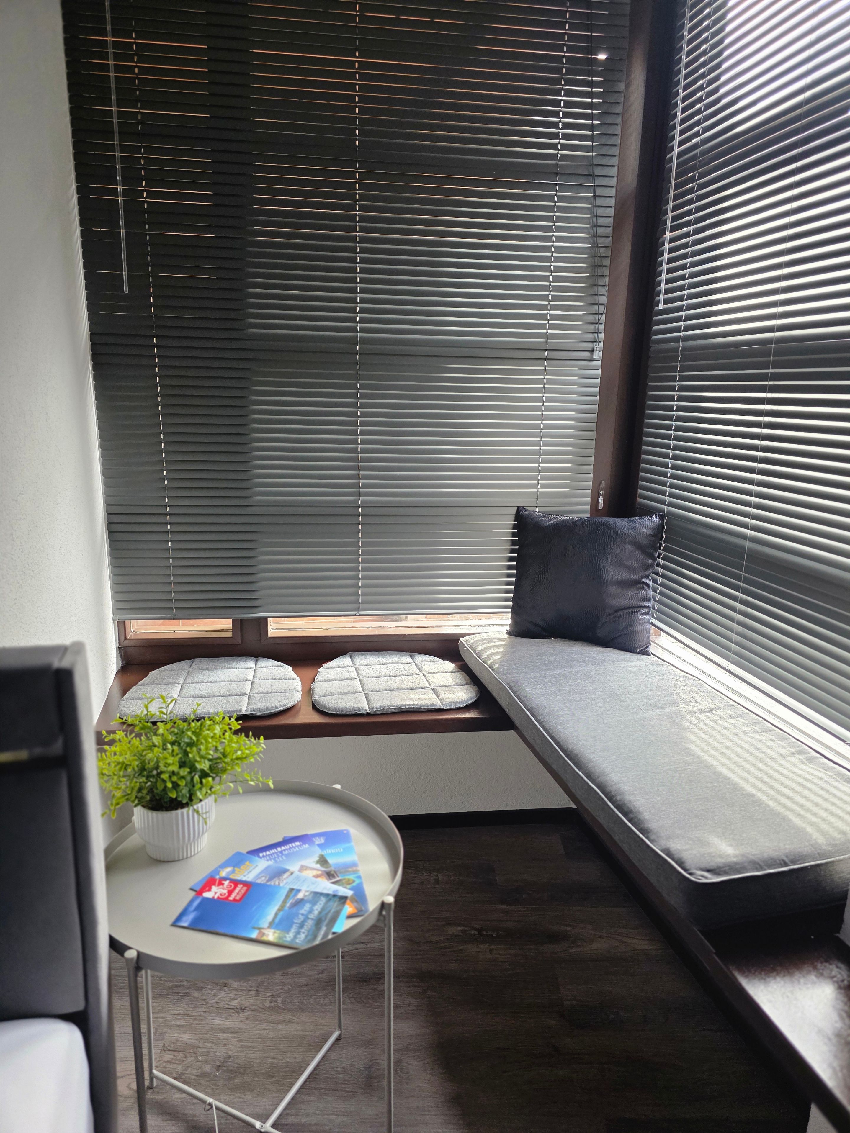 Un rincón con asientos, una ventana, una mesa pequeña con una planta y una revista.