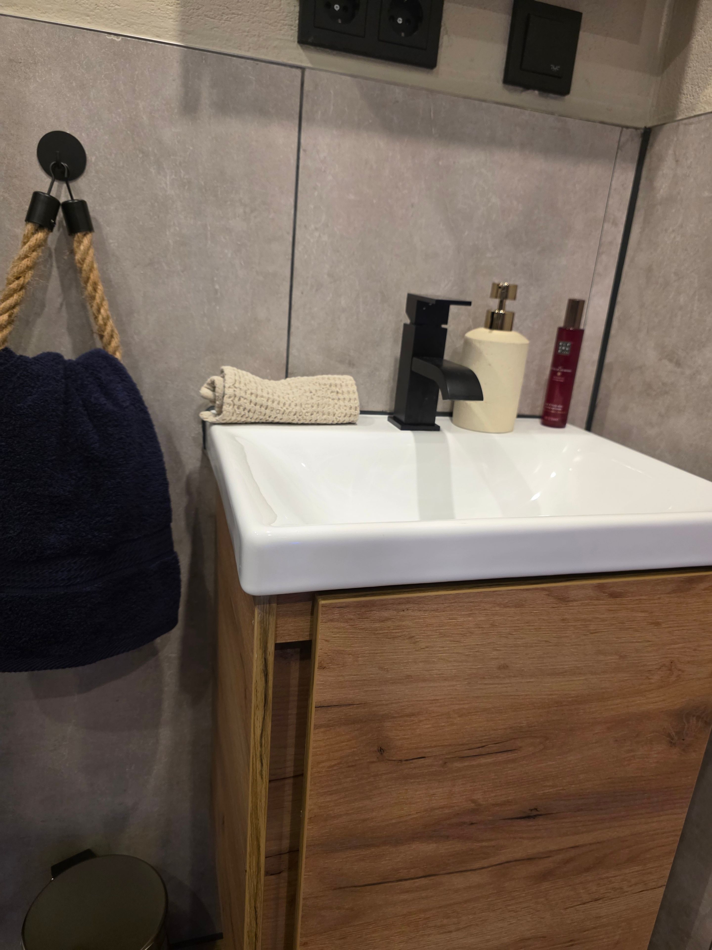 Un baño con lavamanos blanco, mueble de madera, toallero y artículos de aseo.