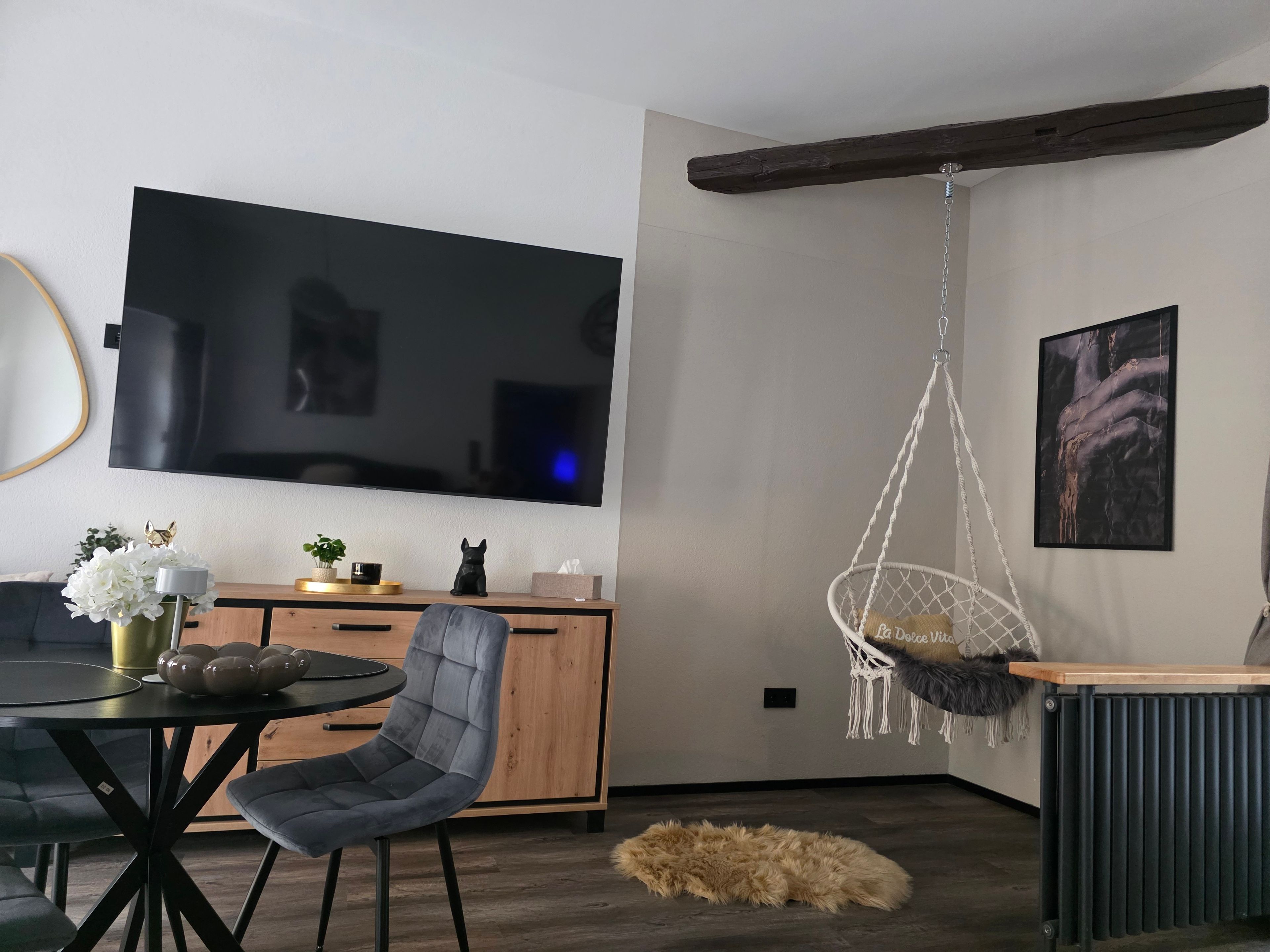 Ein modernes Wohnzimmer mit Flachbildfernseher, Hängesessel, Esstisch mit Stühlen, Sofa und einem dekorativen Teppich.