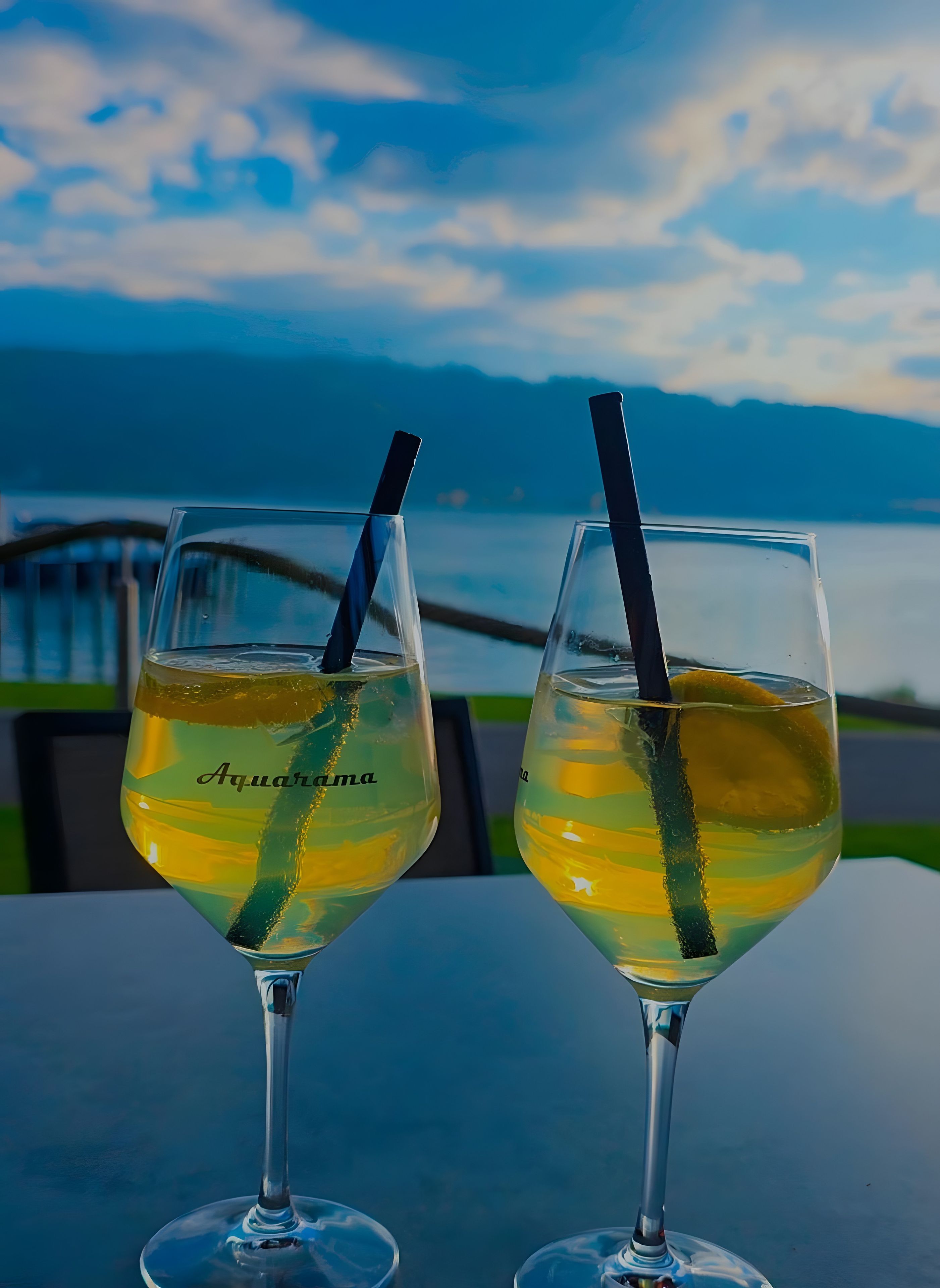 Two glasses of a yellow beverage with a black straw, labeled 'Aquarima,' on a table with a view of a body of water and mountains in the background.