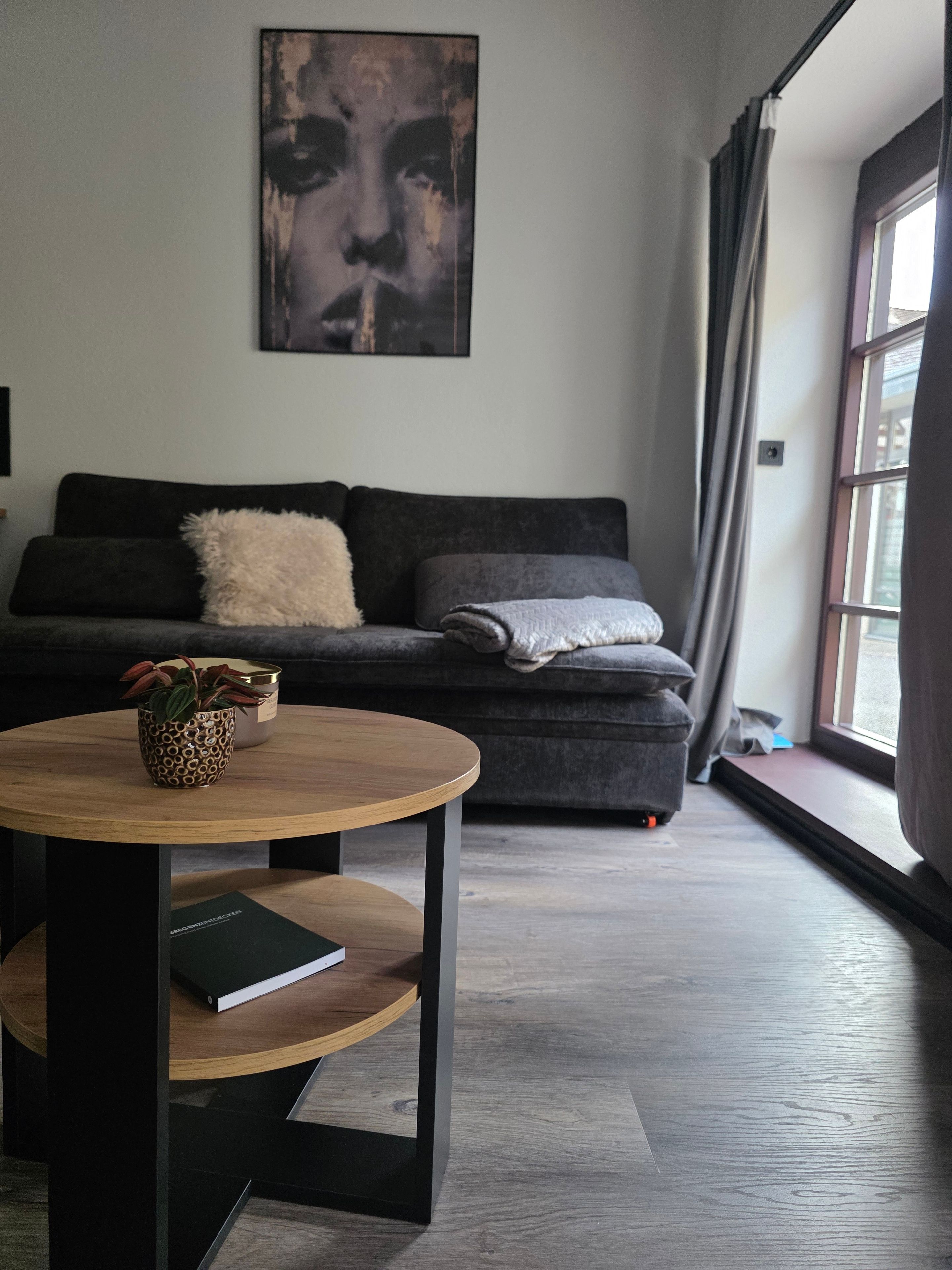 Ein modernes Wohnzimmer mit einem dunklen Sofa, einem runden Holztisch und einem großen Fenster mit Vorhängen.