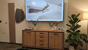 Ein modernes Wohnzimmer mit einem großen Flachbildfernseher an der Wand, einem Holzkasten mit Schubladen, einem schwarzen Teppich und einer üppigen Pflanze im Topf.