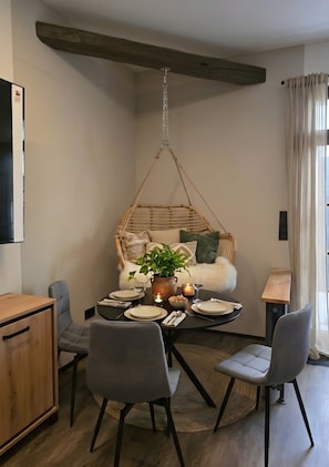 Ein Essbereich mit einem Holztisch, gedeckt für zwei Personen, umgeben von grauen Stühlen. Im Hintergrund ist ein Hängesessel mit Kissen und ein Blumentopf zu sehen.