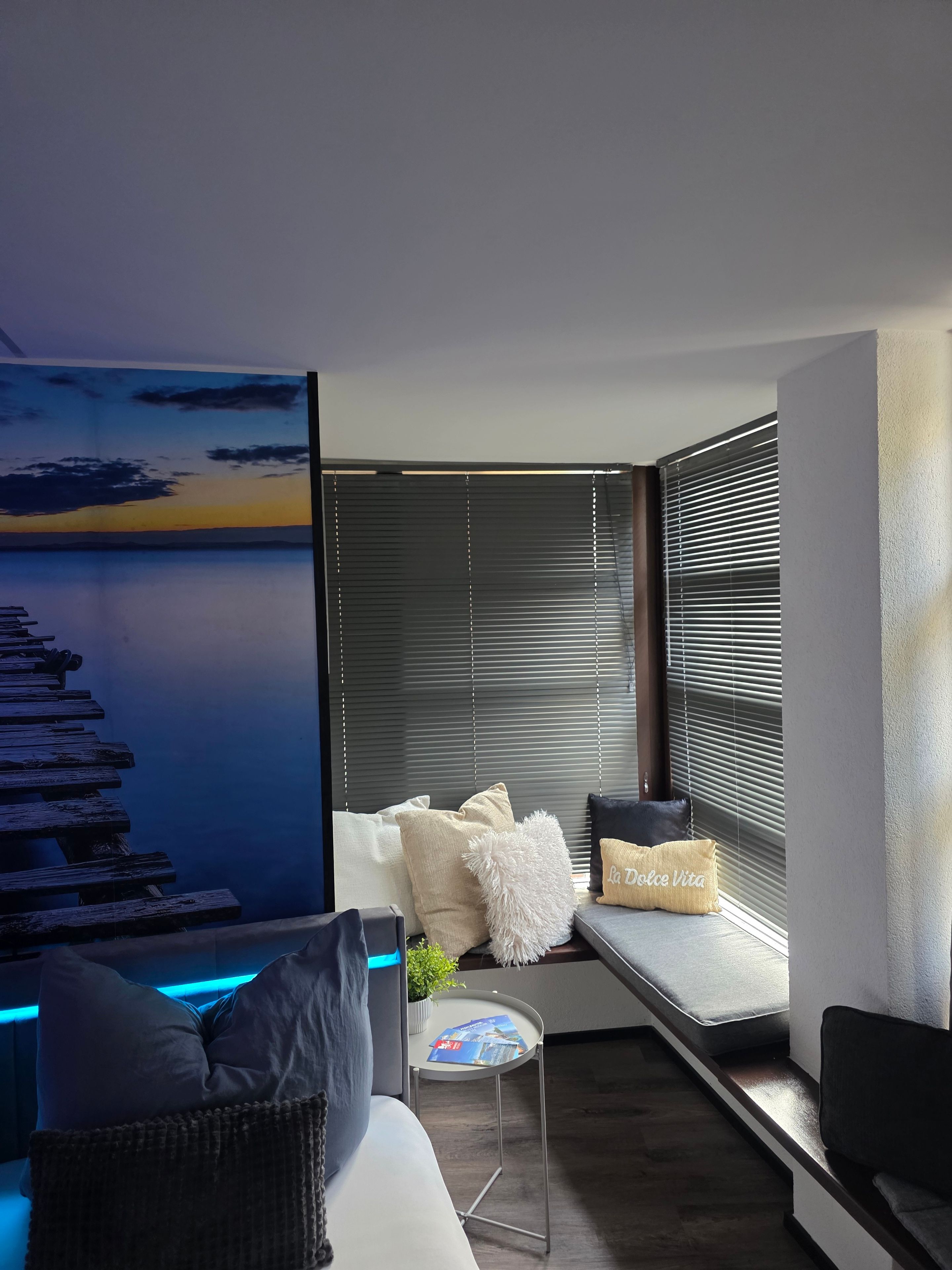 Ein modernes Wohnzimmer mit einer Couch, Kissen, einem kleinen Tisch und einem an der Wand befestigten Fernseher, der eine Sonnenuntergangs-Szene zeigt.