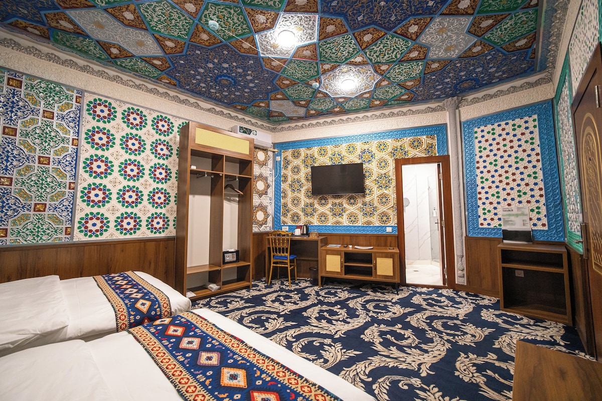 A room with a patterned carpet, a bed with a decorative headboard, a wooden desk, a chair, and a wardrobe.
