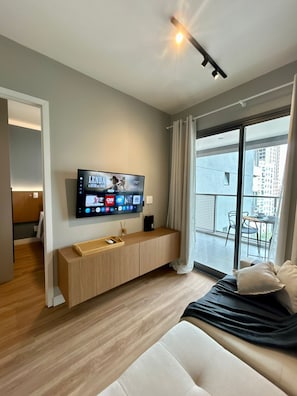 A modern hotel room with a flat-screen TV mounted on the wall, a wooden console table, a sofa, and a balcony with a view of the city. - Level Brooklin 1007 (São Paulo)