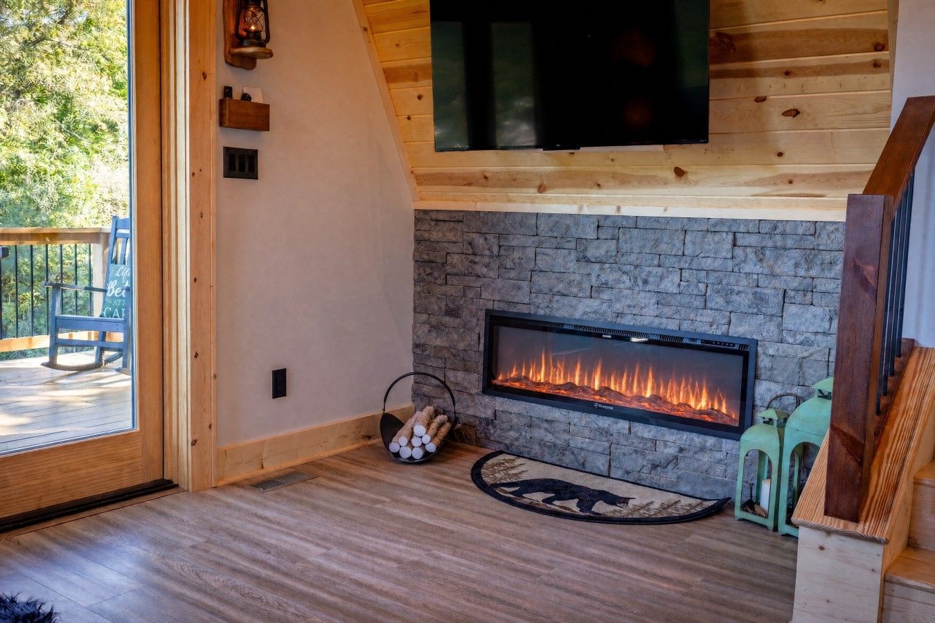 A cozy room with a stone fireplace, a television mounted on the wall, and a wooden deck visible through a glass door.