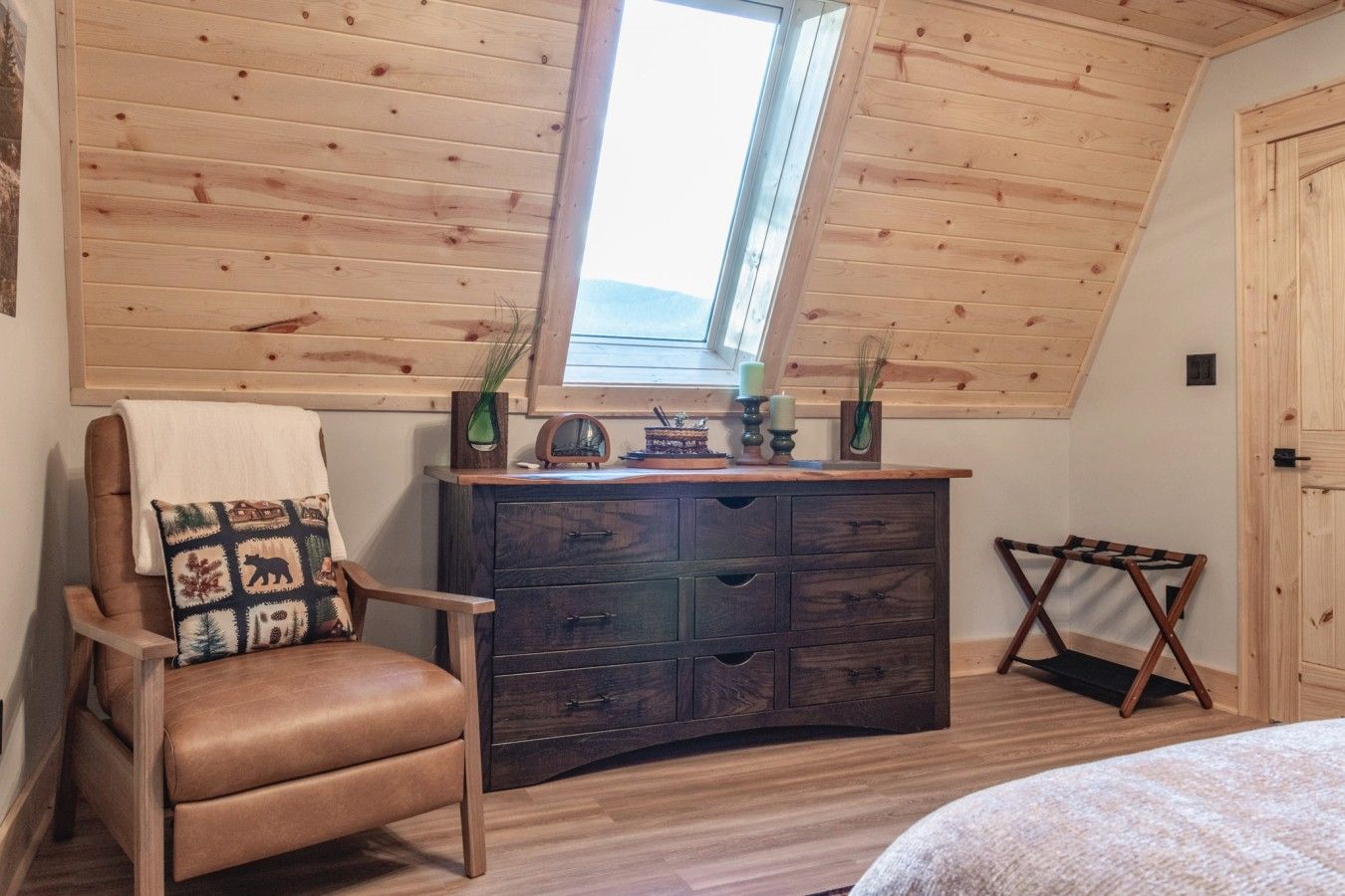 Une pièce chaleureuse avec un plafond en pente, une commode en bois, un fauteuil en cuir et une petite table agrémentée d’objets décoratifs.