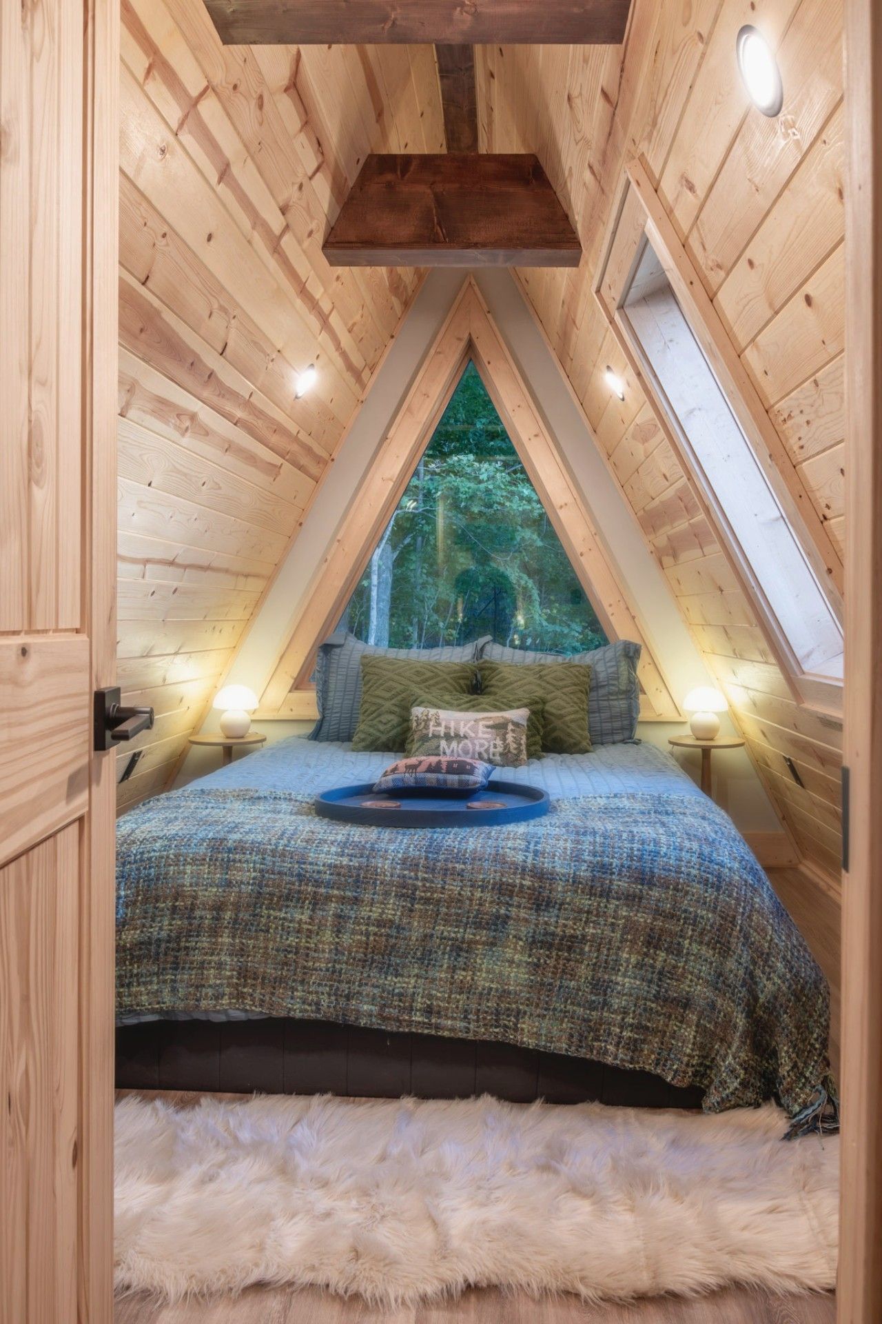 A cozy attic bedroom with a sloped ceiling, a bed with a blue blanket, and a view of the forest through a skylight.