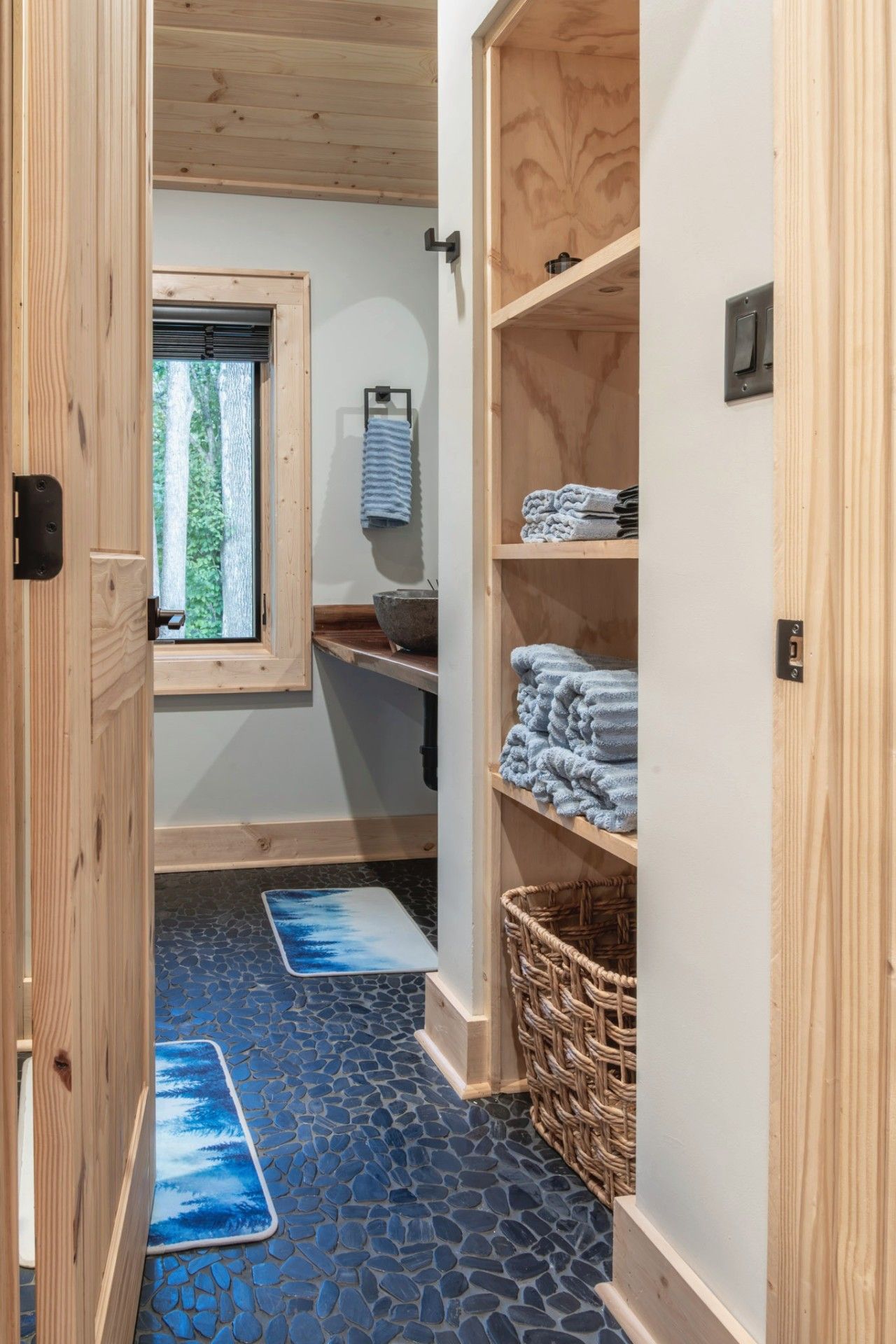 A wooden interior with a stone-tiled floor, a window with a view of trees, and shelves with folded towels.
