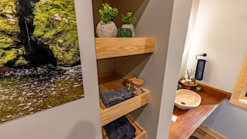 Une salle de bain avec une étagère en bois où sont rangées les serviettes, un lavabo avec un receveur en pierre et un porte-serviettes fixé au mur.