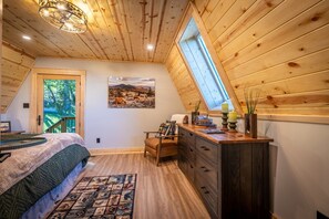 Un dormitorio con techo inclinado, una cama, un aparador, una silla, un ventanal y un cuadro en la pared.