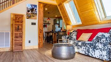 Un salon chaleureux avec un plafond en pente, une porte en bois, un coin repas et une cuisine à l’arrière-plan.