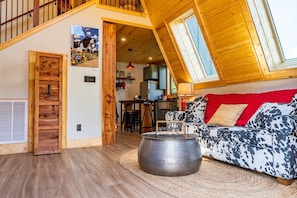 Een gezellige woonkamer met een schuine kap, een houten deur, een eethoek en een keuken op de achtergrond.