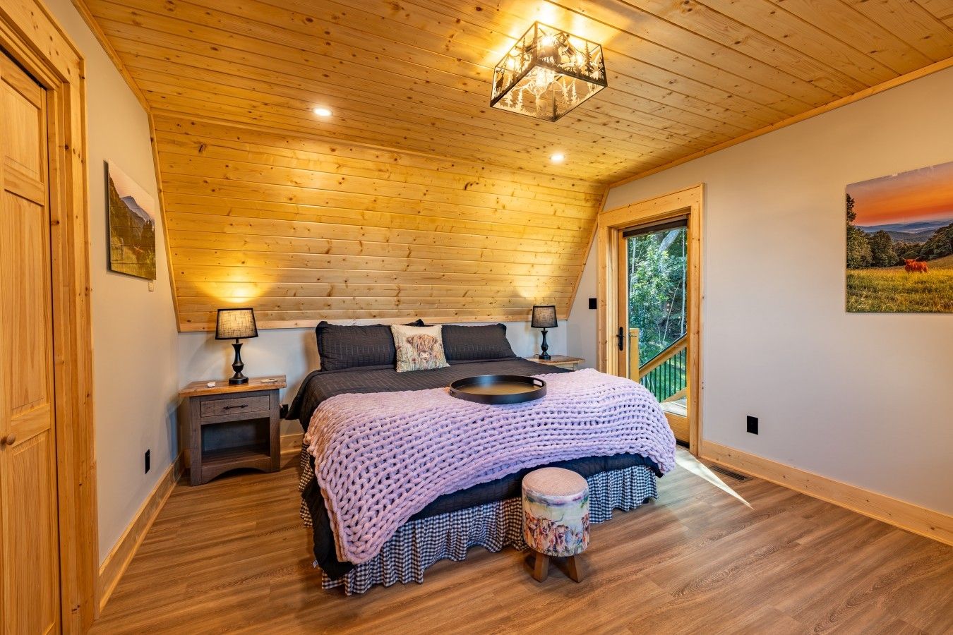 Ein Schlafzimmer mit Holzbalkendecke, einem Bett mit Strickdecke, einem Nachttisch, einer Lampe und Blick ins Freie durch eine Tür.