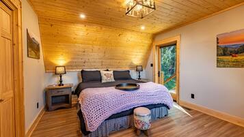 Ein Schlafzimmer mit Holzbalkendecke, einem Bett mit Strickdecke, einem Nachttisch, einer Lampe und Blick ins Freie durch eine Tür.