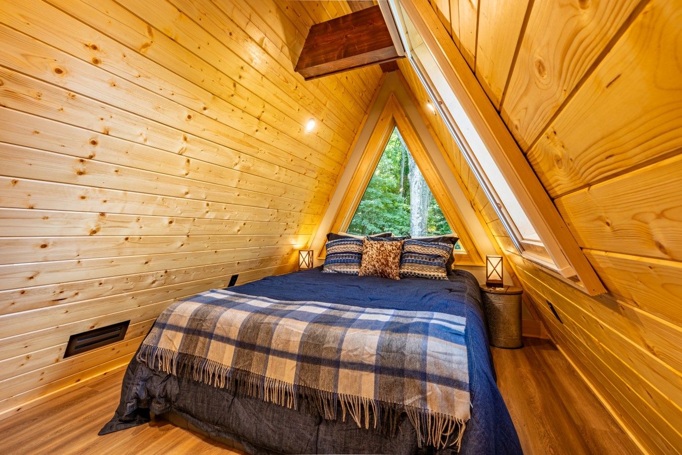 Une pièce chaleureuse aux murs en bois et au plafond en pente, avec un lit recouvert d’une couverture à carreaux et une fenêtre donnant sur un paysage verdoyant.