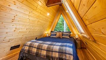 Une pièce chaleureuse aux murs en bois et au plafond en pente, avec un lit recouvert d’une couverture à carreaux et une fenêtre donnant sur un paysage verdoyant.