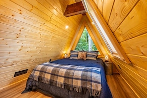 Een knus houten vertrek met schuine wanden, een bed met een geruite deken en een raam met uitzicht op groen.