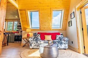 Un accogliente interno di una baita con soffitto in legno, un divano con una coperta rossa, un'area pranzo con tavolo e sedie, e una cucina con elettrodomestici moderni.