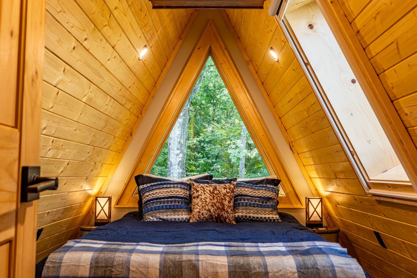 Une chambre mansardée chaleureuse, avec un plafond en pente, un lit recouvert d’une literie à motifs et une vue sur les arbres grâce à une lucarne.