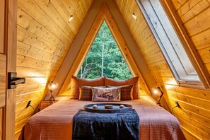 Una camera da letto mansardata accogliente, con soffitto spiovente, un'ampia finestra vista sulla foresta, un letto con copriletto a quadri e due abat-jour.