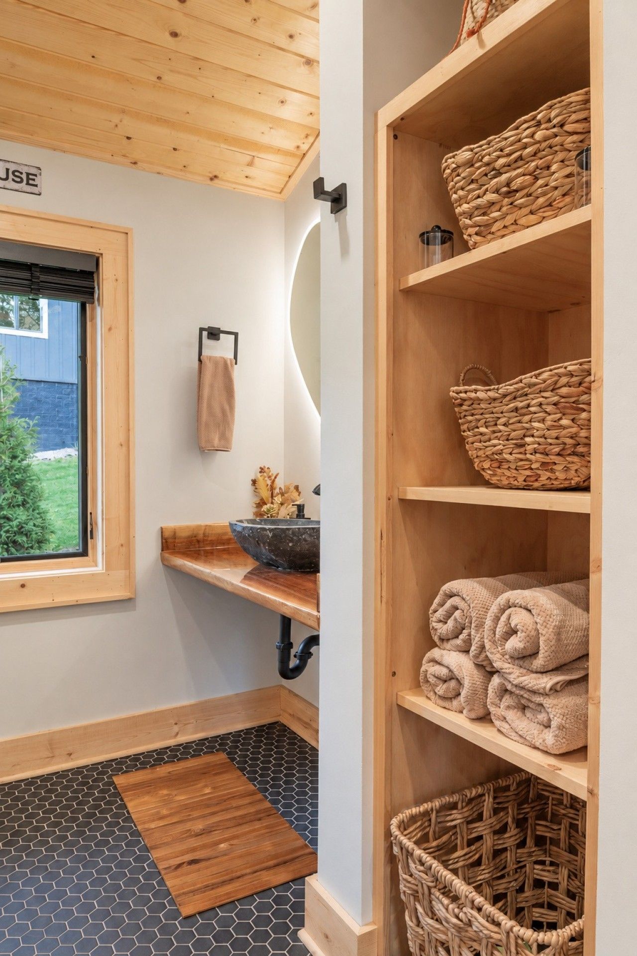 A bathroom with a wooden ceiling, a window, a sink, and shelves with woven baskets and towels.