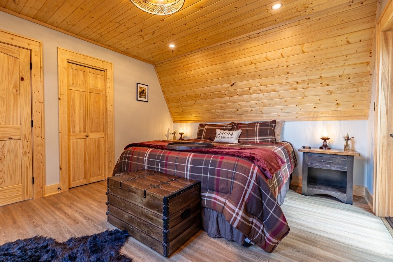 Une chambre avec un plafond en bois, un lit recouvert d’une couette à carreaux, une commode et une table de chevet avec une lampe.